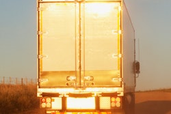 Truck driving down road with sun on its trailer.