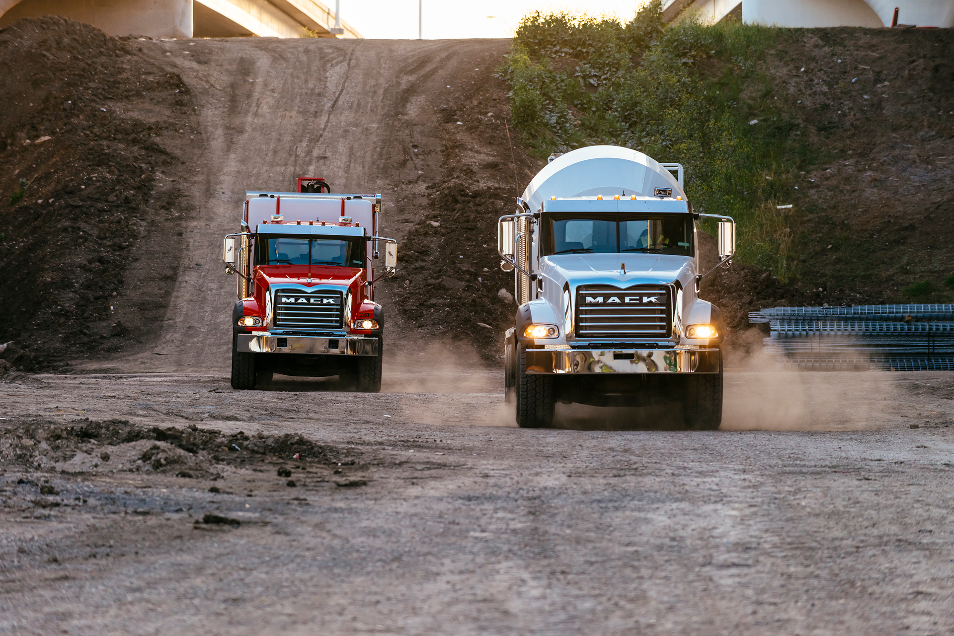 Two Mack Trucks driving down hill on gravel