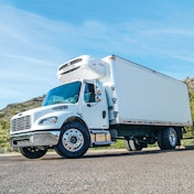Refrigerated box truck parked on pavement in front of a hill.