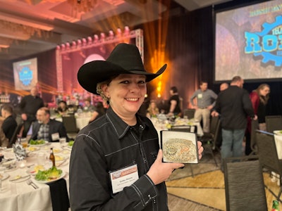 A woman in a black shirt and black cowboy hat holds up a belt buckle.