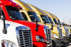 A line of new Class 8 trucks waiting on a lot. From left, they are white, red, and three gold ones.