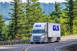 A truck going down the road with pine trees in the background.