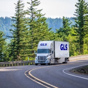 A truck going down the road with pine trees in the background.