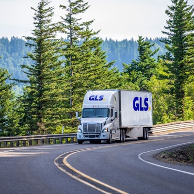 A truck going down the road with pine trees in the background.