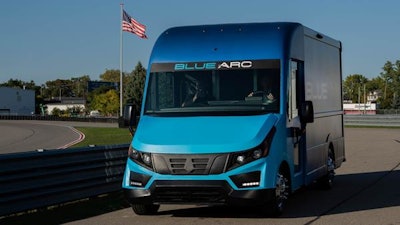 A blue Blue Arc electric delivery truck drives down a road.