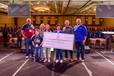 A group of people, some wearing St. Jude and some wearing FleetPride shirts, stand on stage in a crowded ballroom holding a giant check.