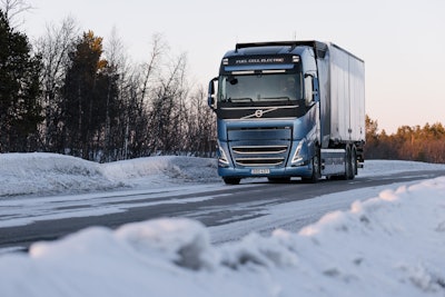 A blue truck going down a snowy road lined with trees.