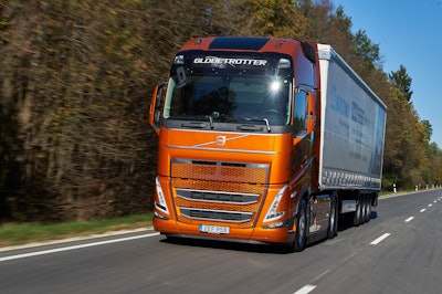 An orange Volvo truck hauling a trailer along a tree-lined road.