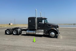 A gray Peterbilt truck without a trailer in front of a wet pad of asphalt.