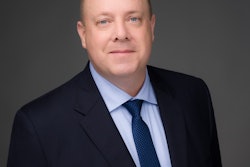 A picture of white man with blue eyes and bald head in a suit against a gray background.