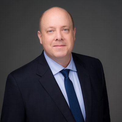 A picture of white man with blue eyes and bald head in a suit against a gray background.