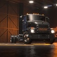 A dark gray electric medium-duty truck in a hangar with a light-up Mack bulldog logo behind it.