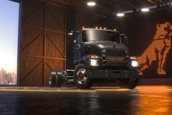 A dark gray electric medium-duty truck in a hangar with a light-up Mack bulldog logo behind it.