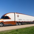An orange and white Class 8 truck by Navistar with a trailer that has logos on it.