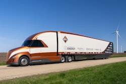 An orange and white Class 8 truck by Navistar with a trailer that has logos on it.