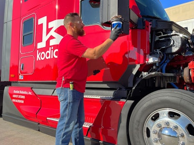 A man in a red shirt and jeans adjusts a part on the side of a bright red Kodiak autonomous Class 8 truck.