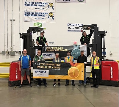 People holding up banners that read '2023 top performing warehouse award' and '2023 most improved warehouse award' with forklifts in the background.