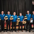 A group of men in blue shirts hold yellow racing helmets in front of a Team Penske car.
