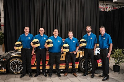 A group of men in blue shirts hold yellow racing helmets in front of a Team Penske car.