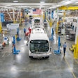 Buses being assembled in a plant with an American flag in the background.