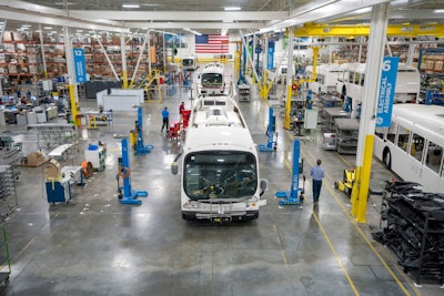 Buses being assembled in a plant with an American flag in the background.