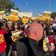 A crowd of picketers holding signs stands in front of a bald man with a bullhorn.