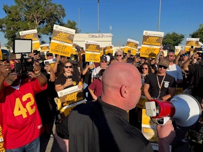 A crowd of picketers holding signs stands in front of a bald man with a bullhorn.
