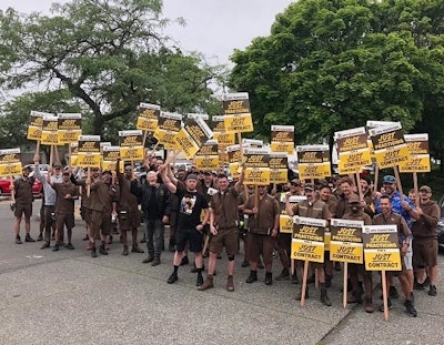 A group of brown-clad UPS workers hold picket signs outdoors.