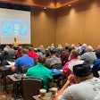 A man in front of a projection screen lectures a room full of people.
