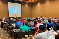 A man in front of a projection screen lectures a room full of people.