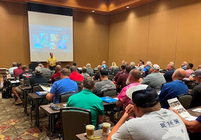 A man in front of a projection screen lectures a room full of people.