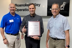 Three men stand in front of a Mack Trucks, Roanoke Valley Operations, sign, the middle one holding a plaque and giving a thumbs up.