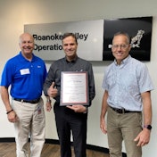Three men stand in front of a Mack Trucks, Roanoke Valley Operations, sign, the middle one holding a plaque and giving a thumbs up.