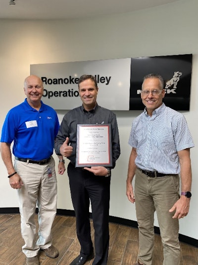 Three men stand in front of a Mack Trucks, Roanoke Valley Operations, sign, the middle one holding a plaque and giving a thumbs up.