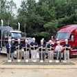 Eight people with shovels stand in front of a box of dirt and two Class 8 trucks.