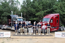 Eight people with shovels stand in front of a box of dirt and two Class 8 trucks.