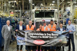 A group of people in suits and orange safety vests stand in front of a truck. They are holding a banner with the Peterbilt logo and the Massey Motor Freight Logo. It reads, 'Congratulations to Troy Massey, recipient of the 100,00th Model 389'