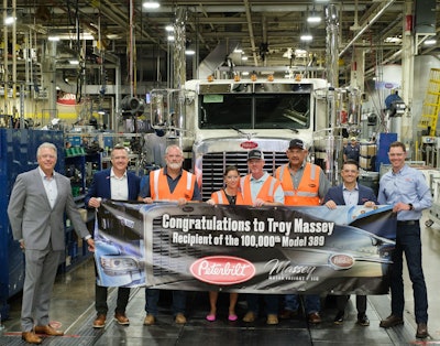 A group of people in suits and orange safety vests stand in front of a truck. They are holding a banner with the Peterbilt logo and the Massey Motor Freight Logo. It reads, 'Congratulations to Troy Massey, recipient of the 100,00th Model 389'