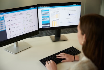 A woman sits at a desk looking at two computer screens with fleet data displayed by TracKing Pro telematics.