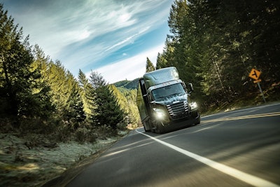 An electric Class 8 truck drives down a road through a forest.