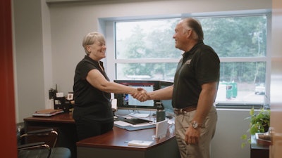 A woman standing behind a desk and a man standing in front of it shake hands.