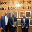 Three men stand in front of a wall of plaques that reads 'American Trucking and Industry Leader Hall of Fame'