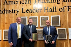 Three men stand in front of a wall of plaques that reads 'American Trucking and Industry Leader Hall of Fame'