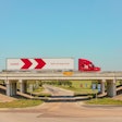 A white-and-red truck and trailer crosses an overpass under a blue sky.