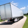 A truck pulling a Utility dry van trailer down a road surrounded by green trees and grass.