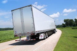 A truck pulling a Utility dry van trailer down a road surrounded by green trees and grass.