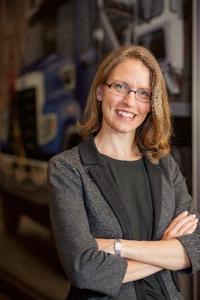 A woman with shoulder-length blonde hair and glasses, with her arms crossed in front of a picture of a truck.