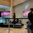 Two men stand on stage in front of a television while a line of other contestants watch.