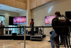 Two men stand on stage in front of a television while a line of other contestants watch.