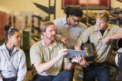 A group of students gather around an instructor holding a tablet in a workshop.
